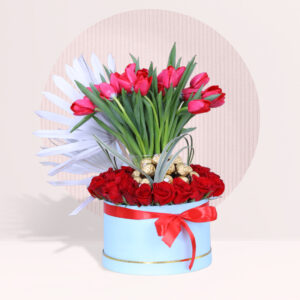A floral arrangement is presented in a light blue, cylindrical box against a light background with a subtle circular pattern. The arrangement features a mix of red tulips, red roses, white palm leaves, and golden chocolates.