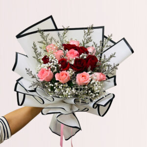a hand holding a bouquet of roses and baby's breath, wrapped in white paper with black edges and tied with a pink ribbon